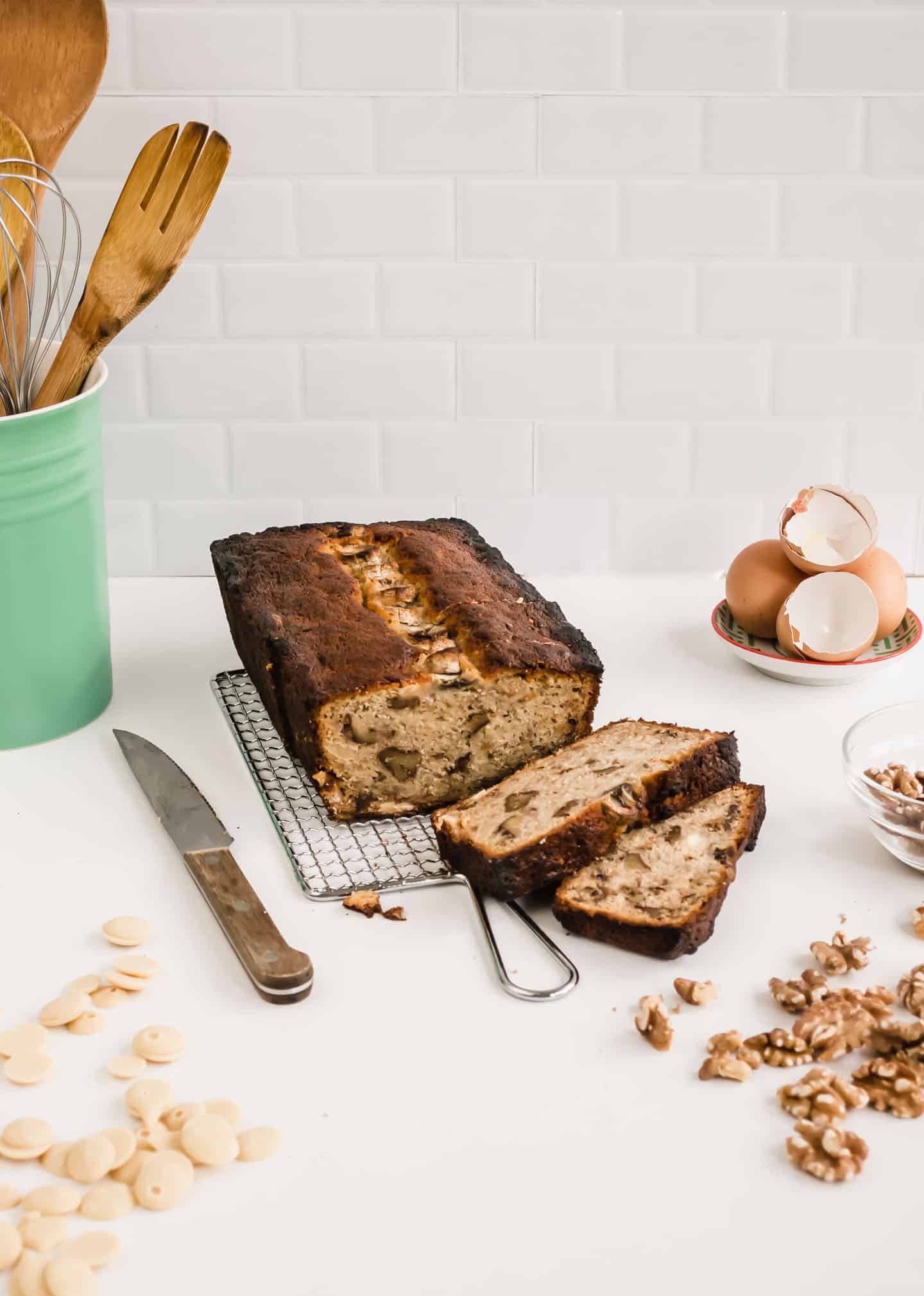 Banana Bread on a cooling rack, a knife next to it, some walnuts and white chocolate chips around it, and some egg shells behind it