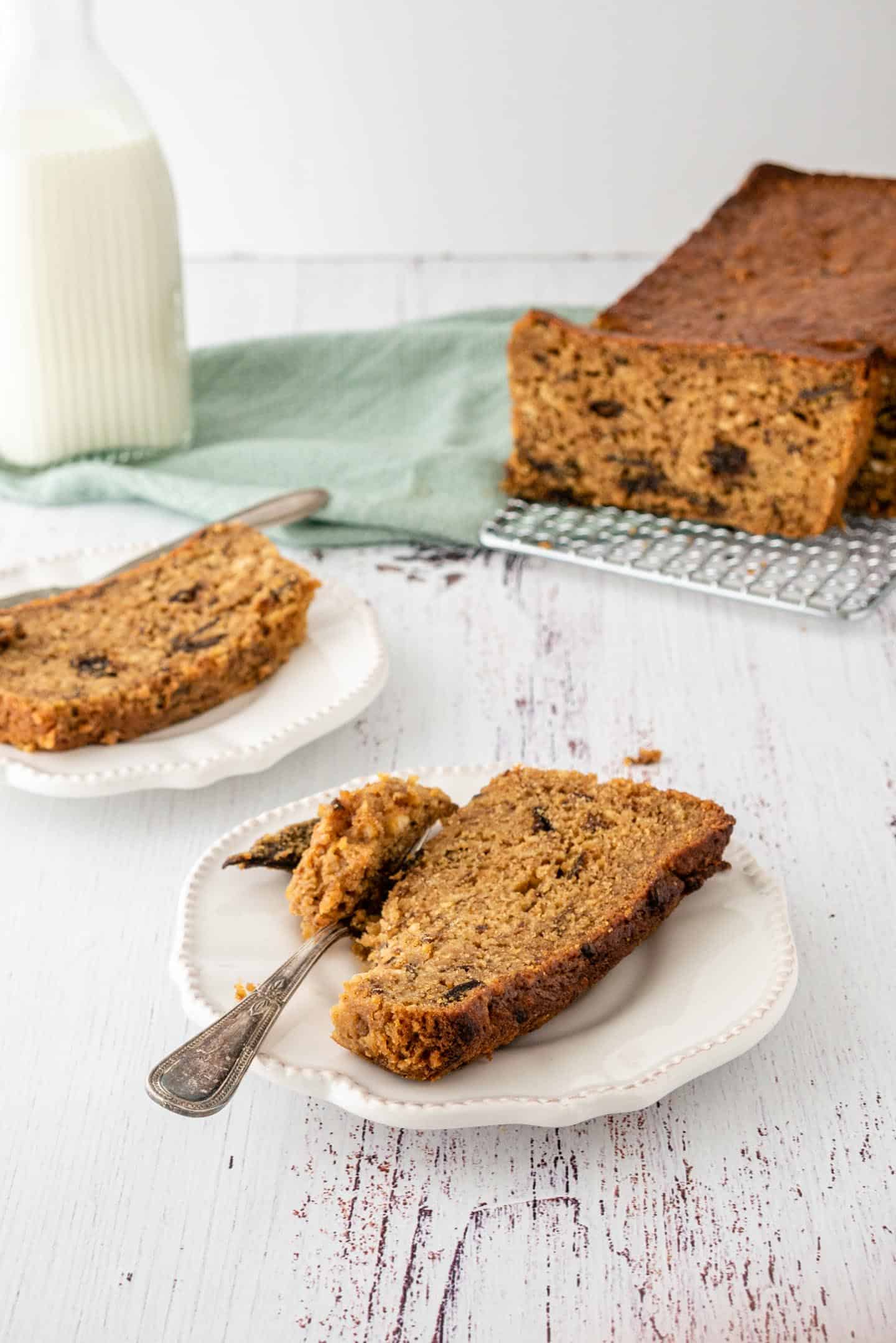 Two slices of cake in front of the whole prune loaf cake on a cooling rack on a corner, a bottle of milk on the other and a green tea towel at the opposite corner.