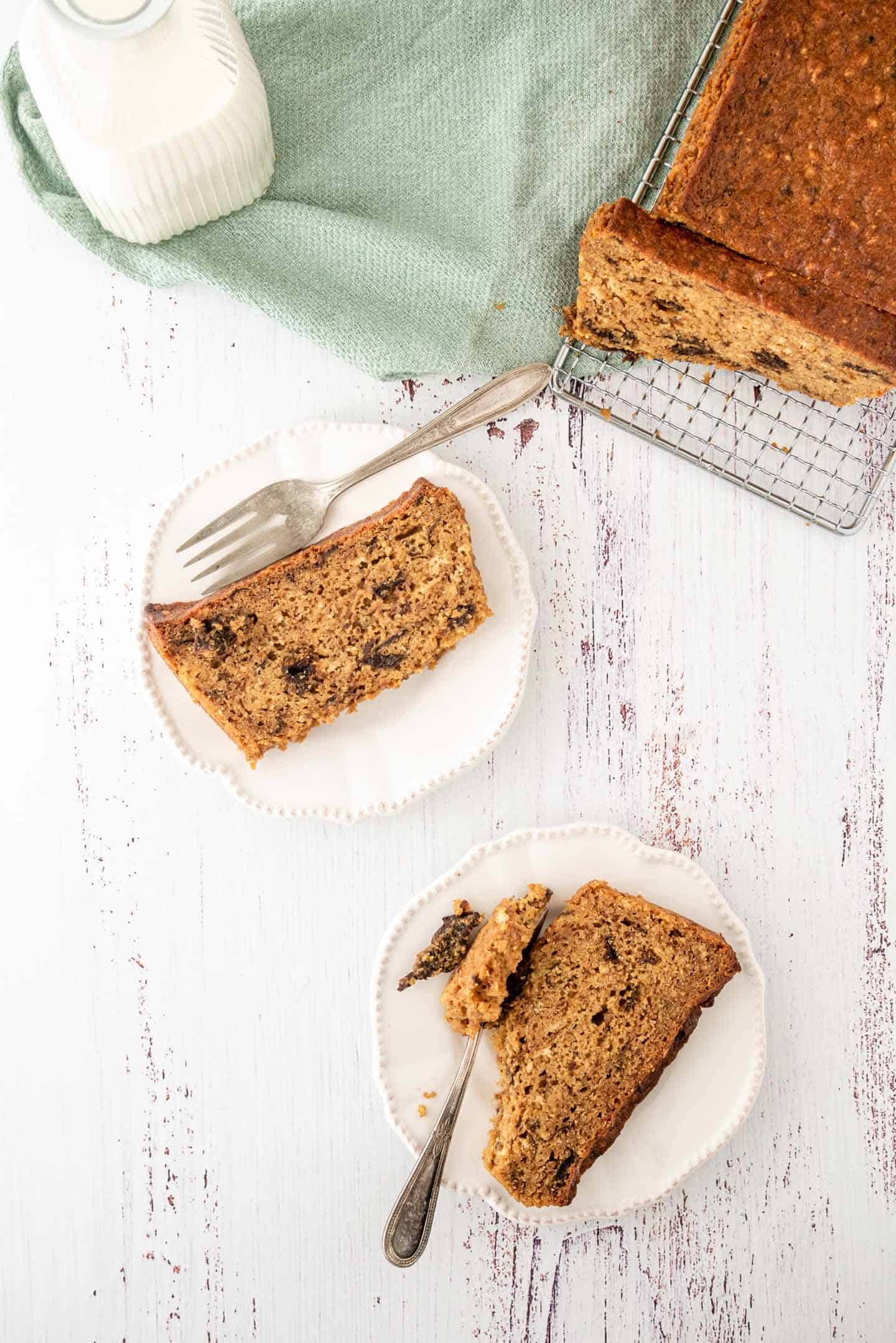 Two slices of cake on white plates, with the whole prune lof cake on a cooling rack in one corner and a green tea towel on the other