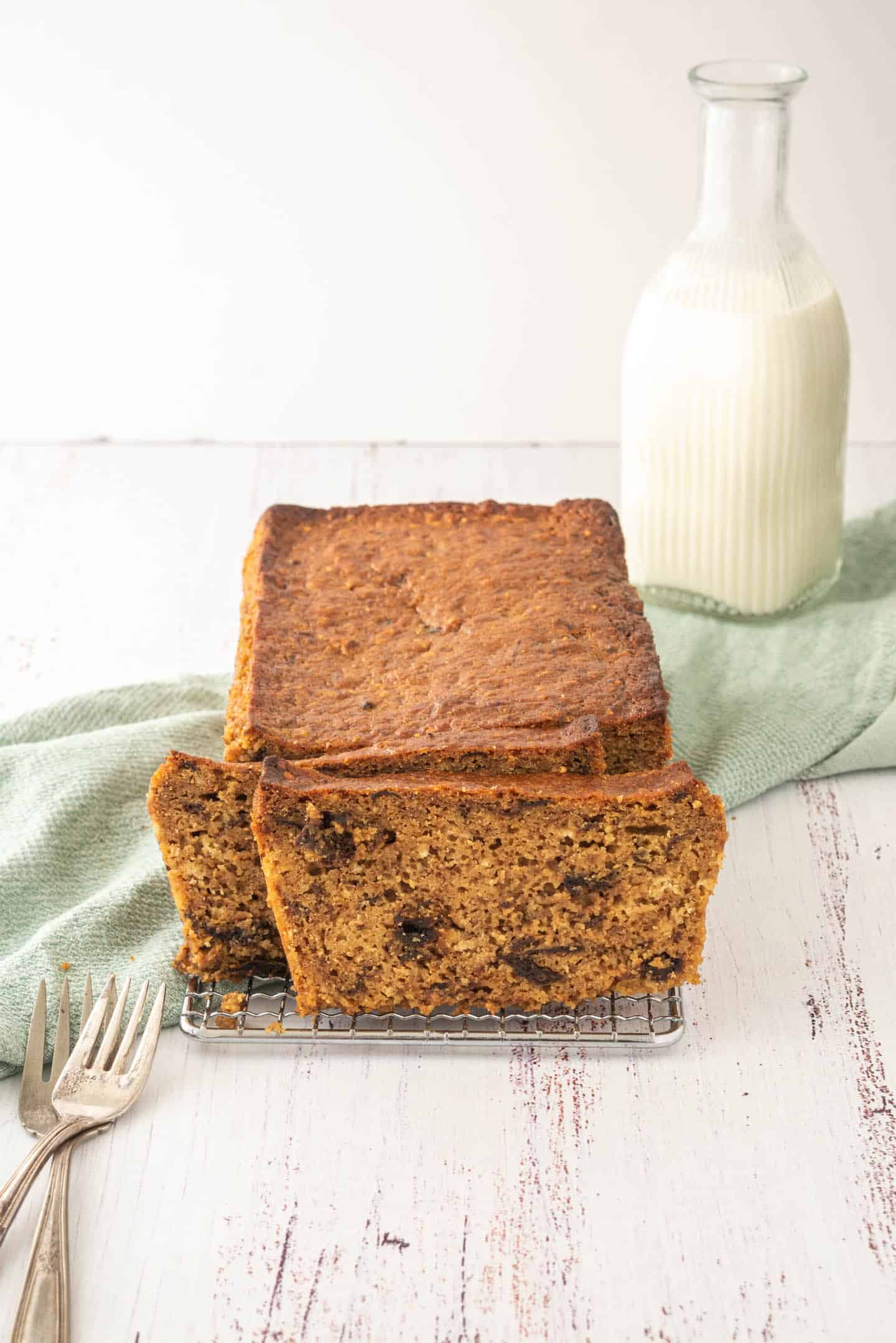 Whole prune cake with some slices on a cooling rack, a bottle of milk in the background and two forks in the left bottom corner