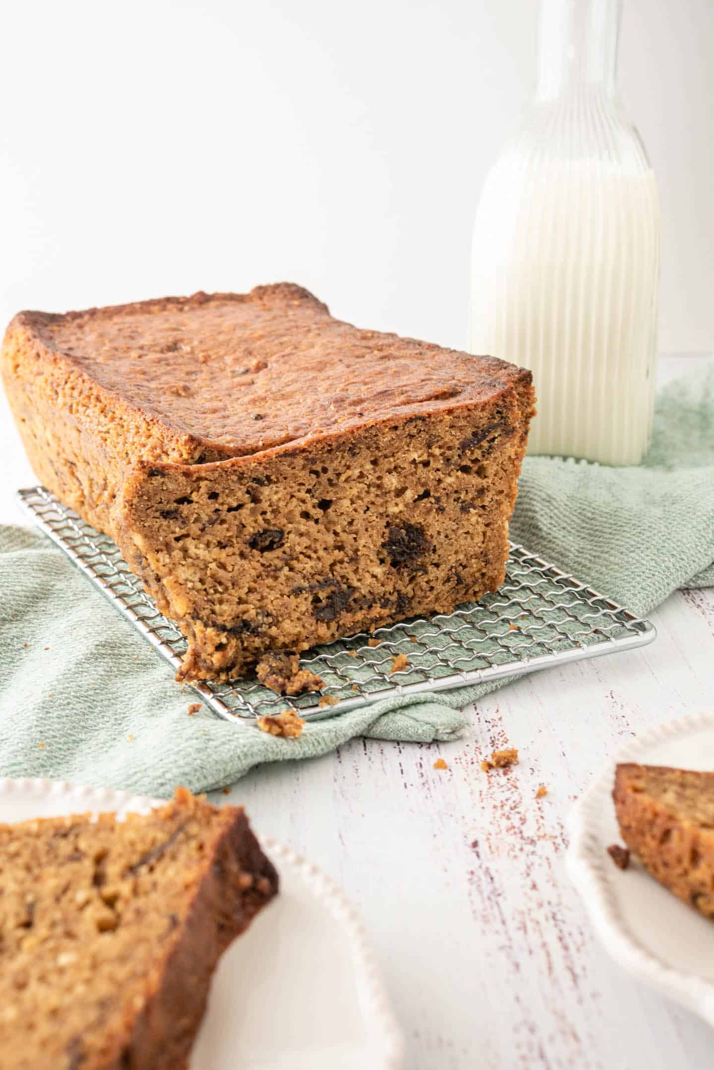 Prune Loaf Cake on a cooling rack and a green tea towel, with a bottle of milk and some slices already cut