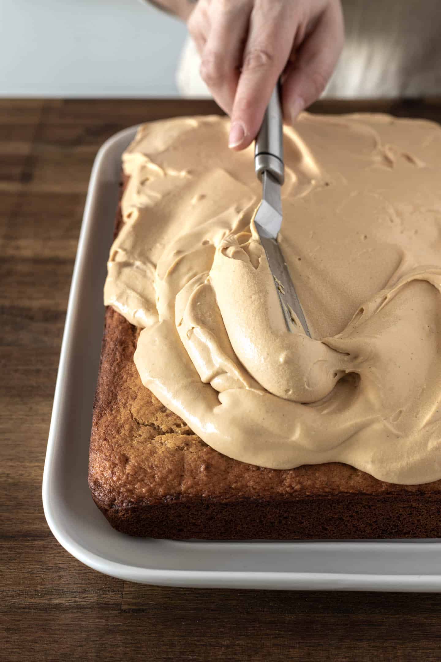 Spatula spreading some dulce de leche buttercream on a banana sheet cake on a white serving plate