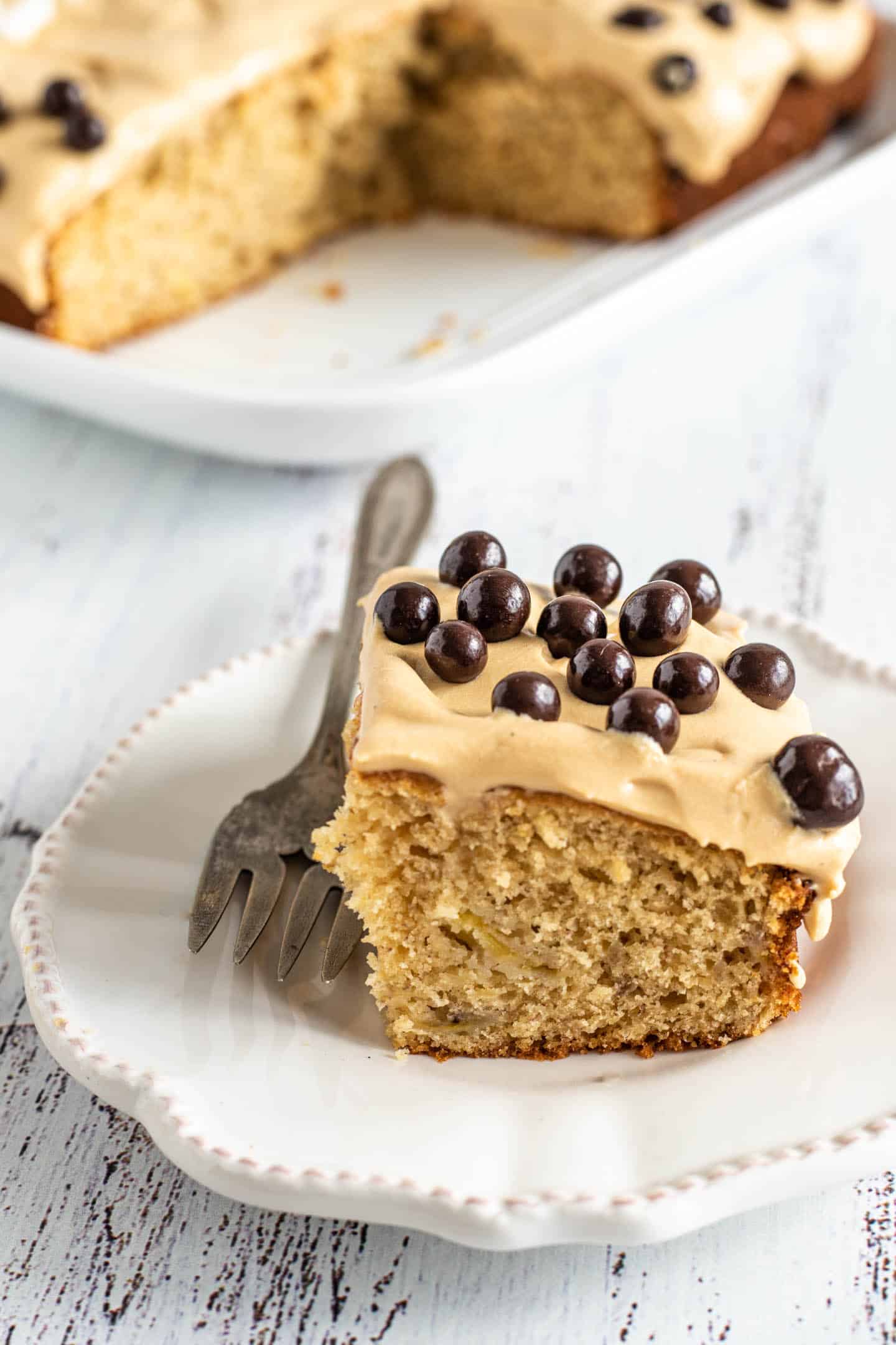 Slice of banana sheet cake on a white plate and a fork, with dulce de leche buttercream and chocolate-covered blueberries