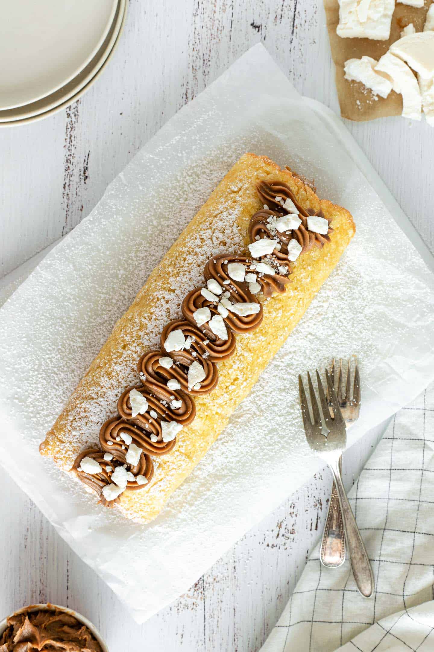 Cake roll overhead shot with dulce de leche and crushed meringues decorations on a white baking sheet, two forks and a small bowl with dulce de leche