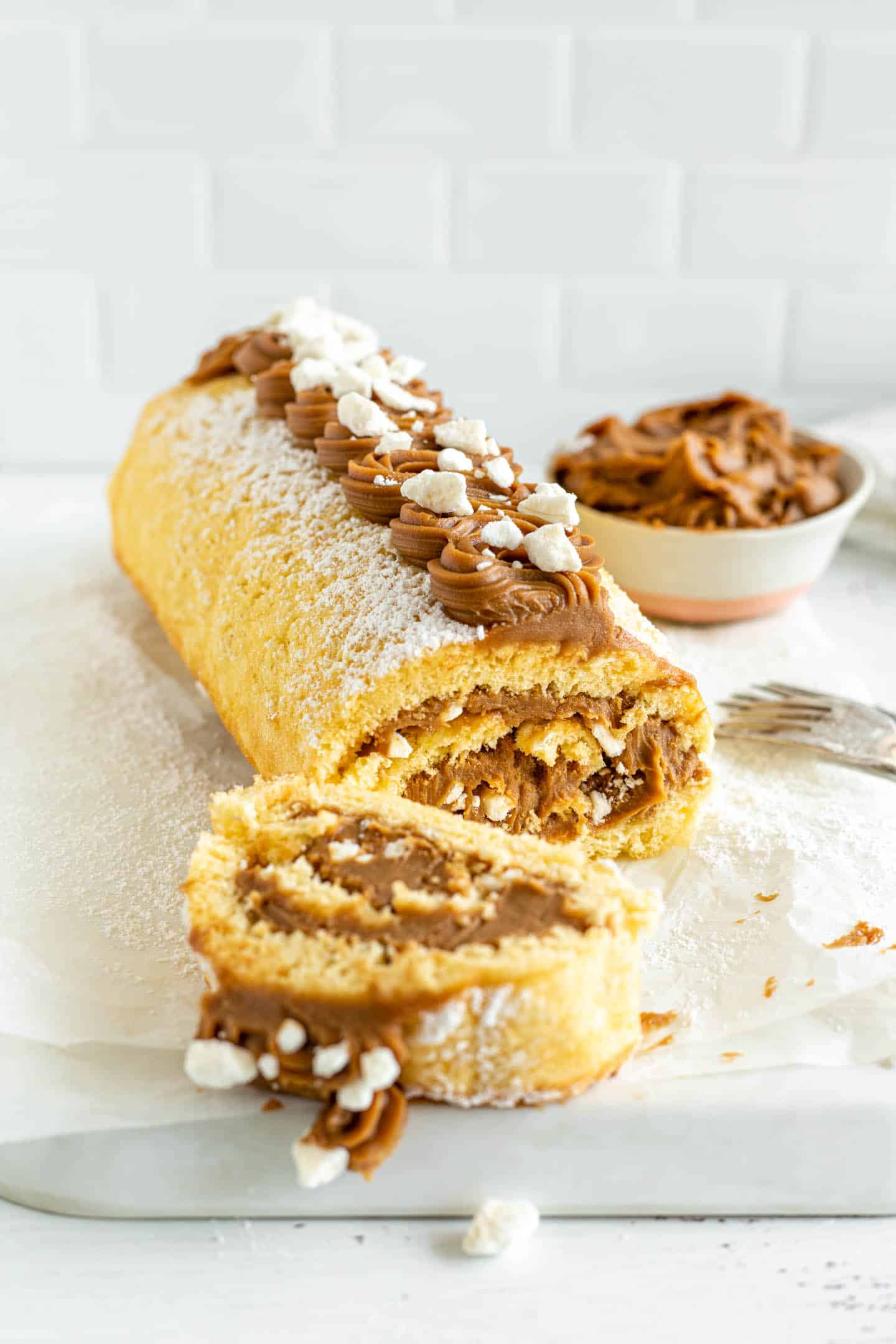Dulce de leche cake roll on a baking sheet, and a small bowl with dulce de leche at the back