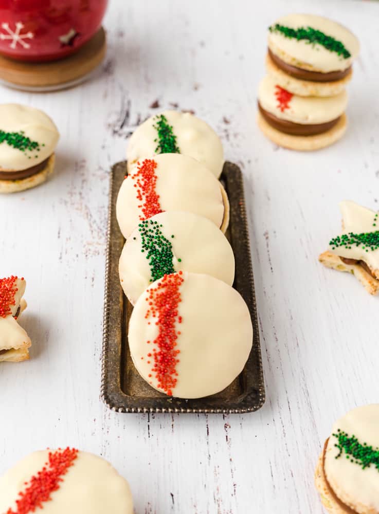 Alfajores cookies covered in white chocolate and red and green sprinkles on a silver small dish, on a white surface