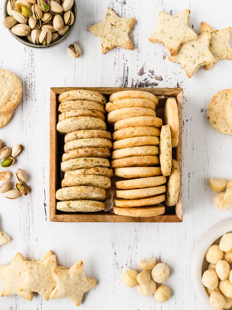 macadamia and pistachios cookies on a wooden tray, on a white surface with some dry nuts around them