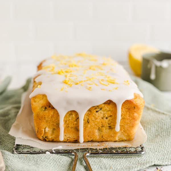 Close up of a lemon zucchini cake on a wire rack, a little green saucer and a lemon on the background, on a green tea towel covered with lemon glaze drips and lemon zest on top