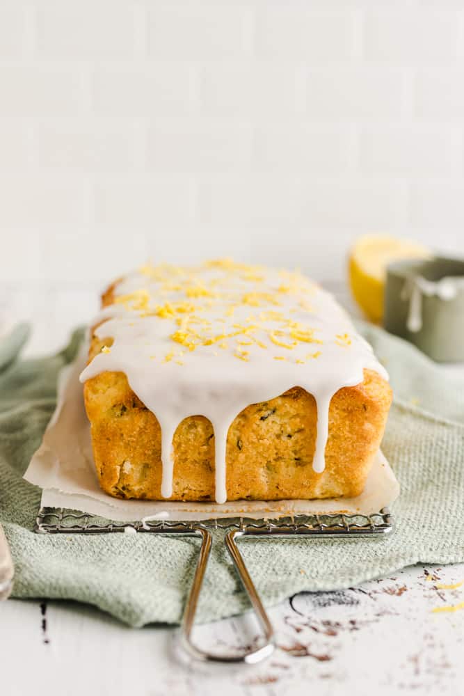 Close up of a lemon zucchini cake on a wire rack, a little green saucer and a lemon on the background, on a green tea towel covered with lemon glaze drips and lemon zest on top