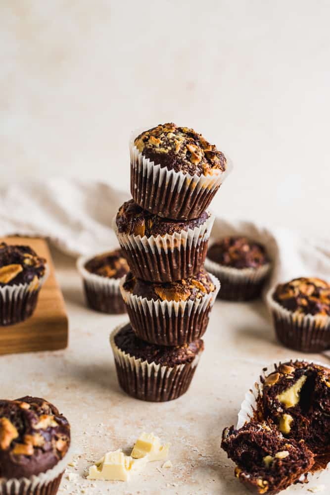 Four stacked double chocolate banana muffins and some scattered muffins, a wooden chopping board and a white tea towel